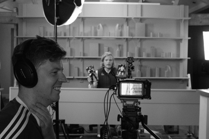 B&W behind-the-scenes: shot of smiling man with headphones. Another man sits at a table, spotlight on him, looking to camera holding up a toy.