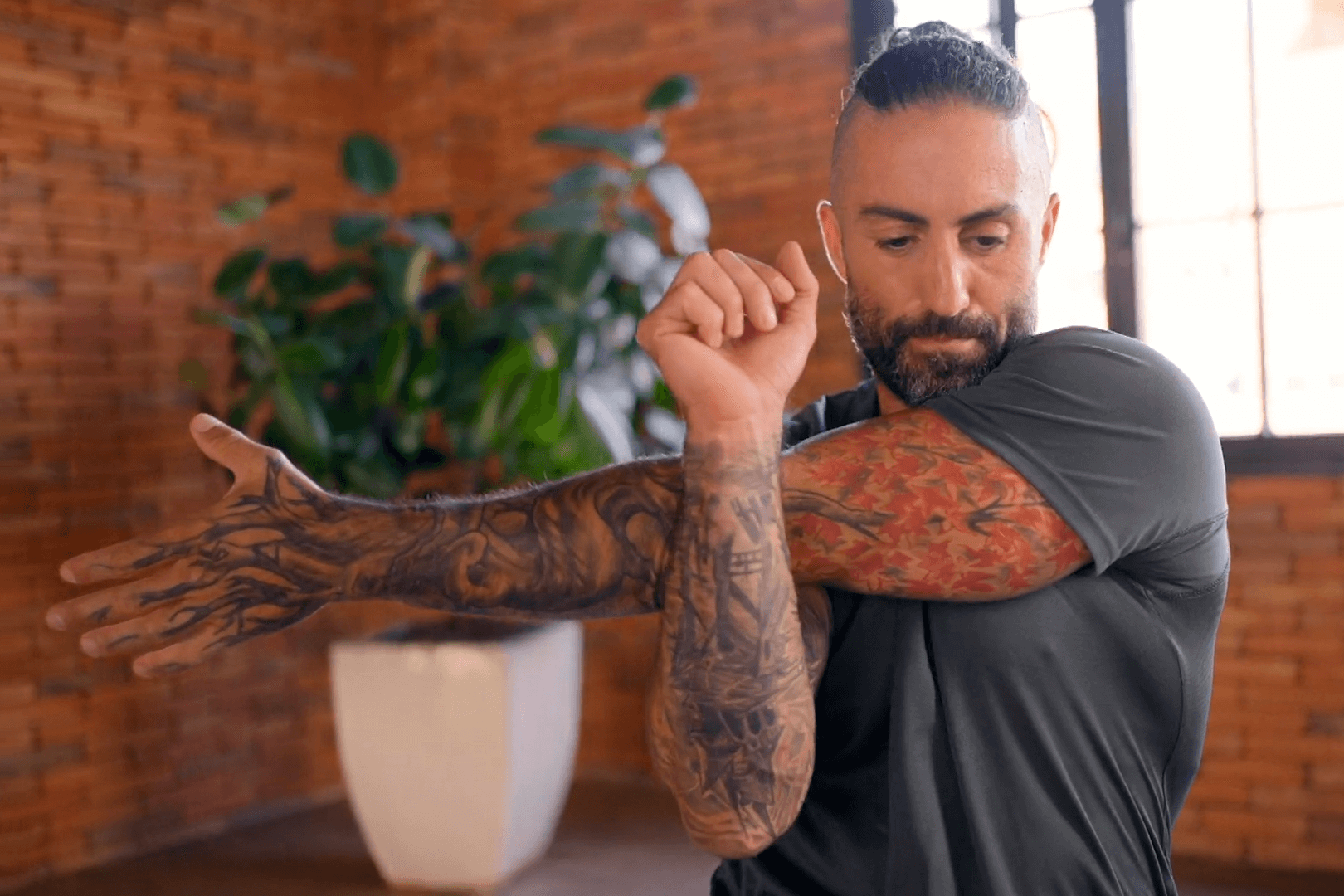 In a daylit room, a man with tattoos covering his arms stretches one arm over the other while wearing a neatly tied back hair and a beard. The room has a brick wall and a big plant pot in the background is blurred.