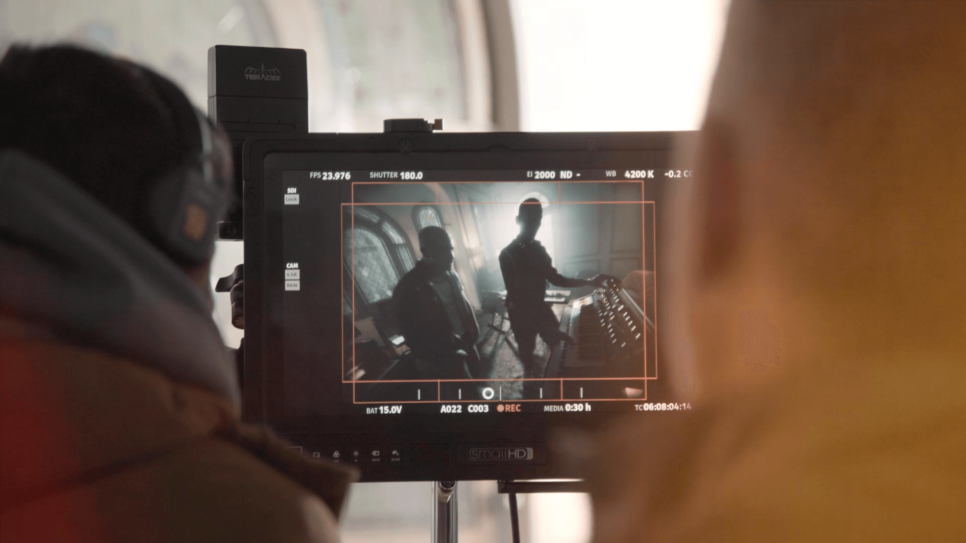 The scene is fairly dark and moody. A man wearing headphones is focused on a screen displaying numbers and other information from a film camera. In front of him, two young men stand before a keyboard, as they are being recorded. A small stream of light is visible in the background.