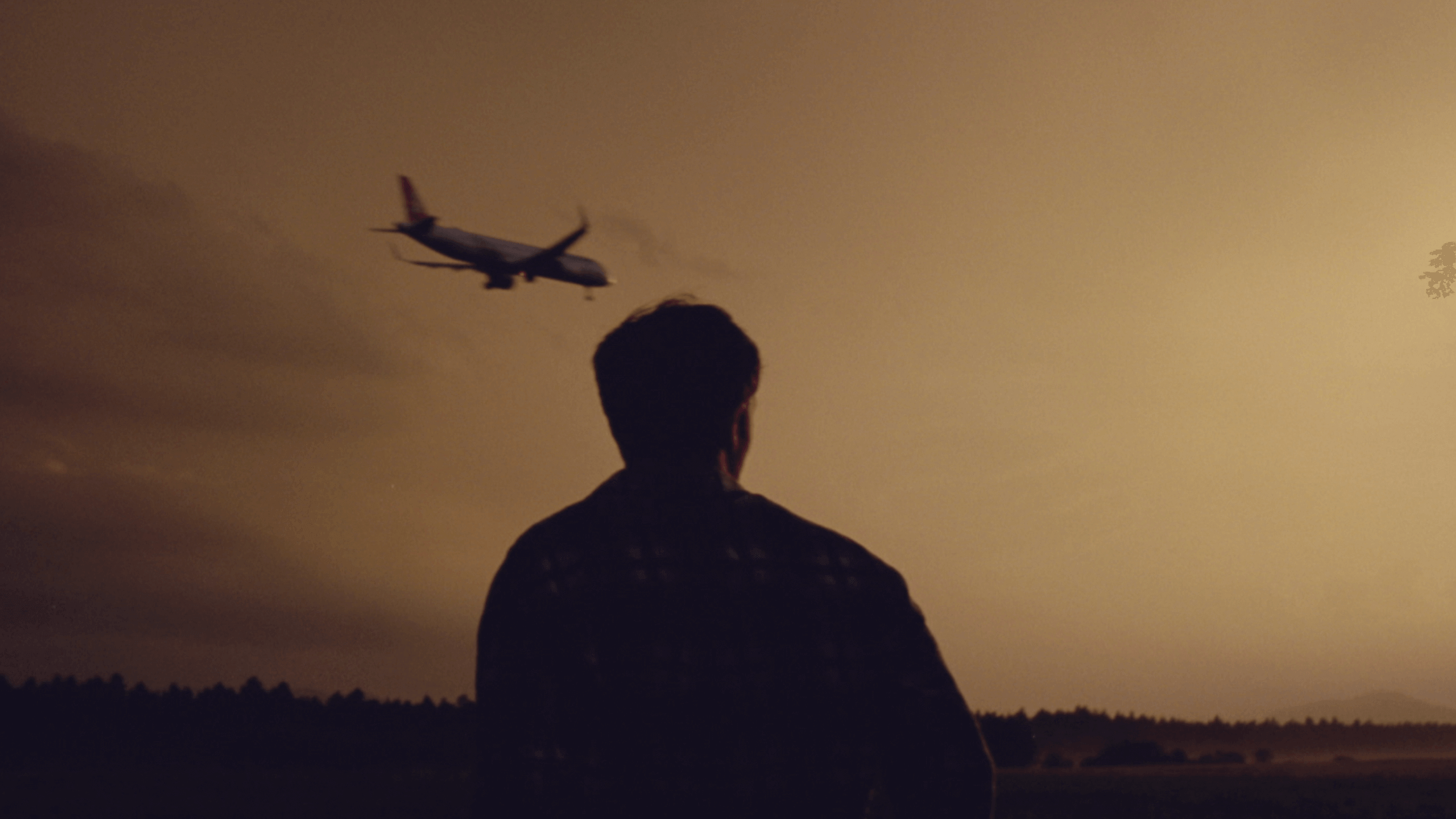 The back of a man standing with a plane in the background approaching for landing during sunset/dusk, creating a thoughtful scene.