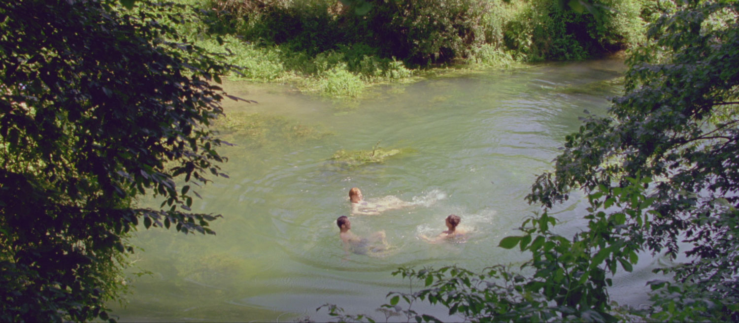 Three individuals are seen swimming in an outdoor pond surrounded by a lush landscape of bushes and trees.