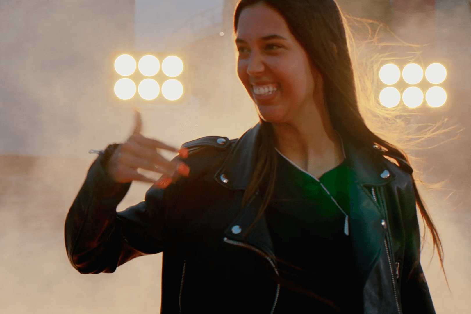 A young Spanish woman with long black hair wears a black leather jacket and smiles, her bright white teeth standing out against the backdrop of big spotlights. She appears energized and ecstatic.