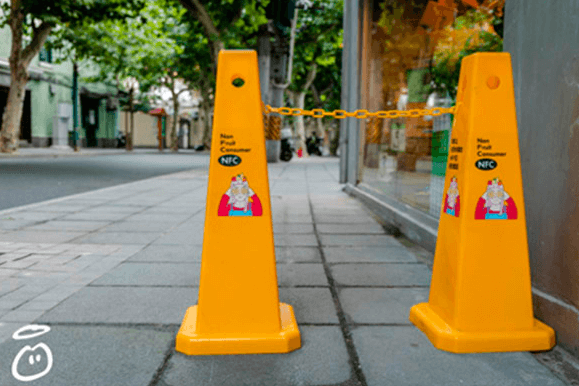 Two bright yellow traffic cones are visible on a city street, adorned with a funky pink sticker. Above, we see the letters 