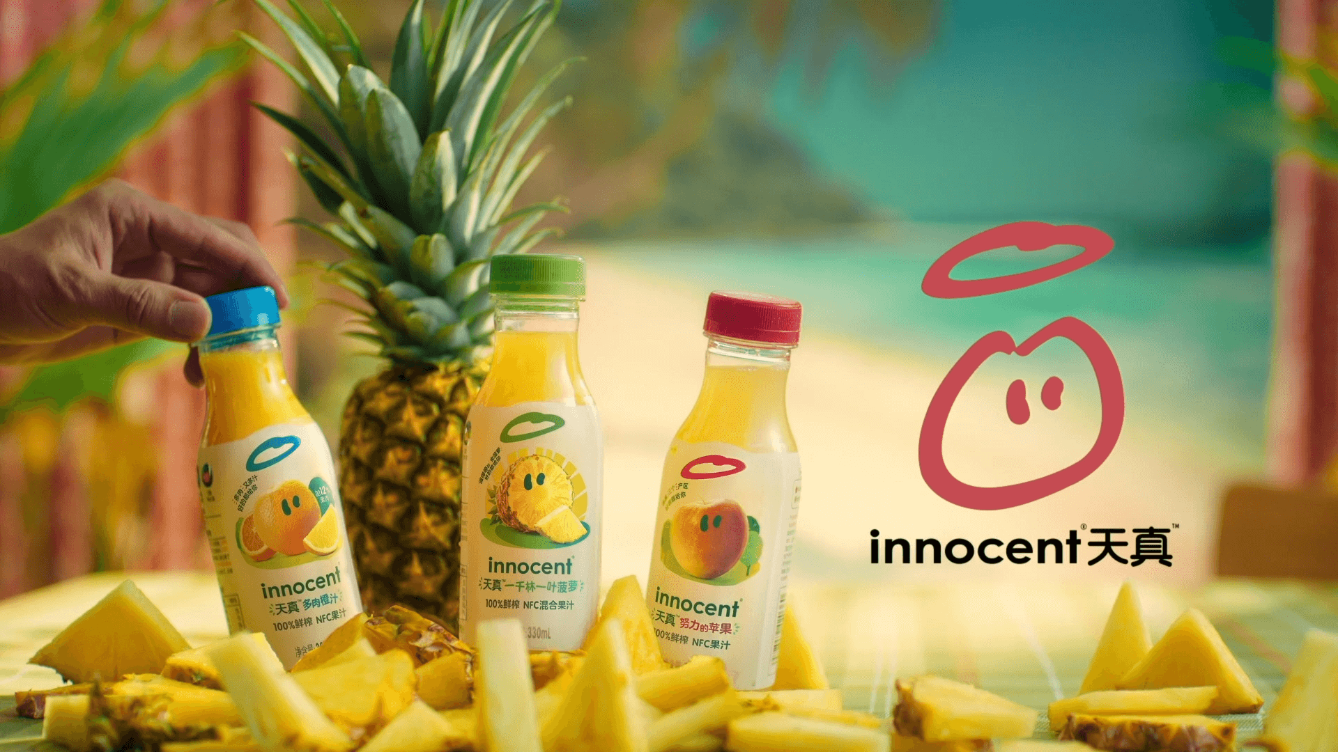 A bright and colorful image of three bottles of Innocent juice, with cut-up pineapples in the foreground and a whole pineapple in the background. A man's hand enters the frame to open one of the bottles. The Innocent logo is prominently displayed on the right.