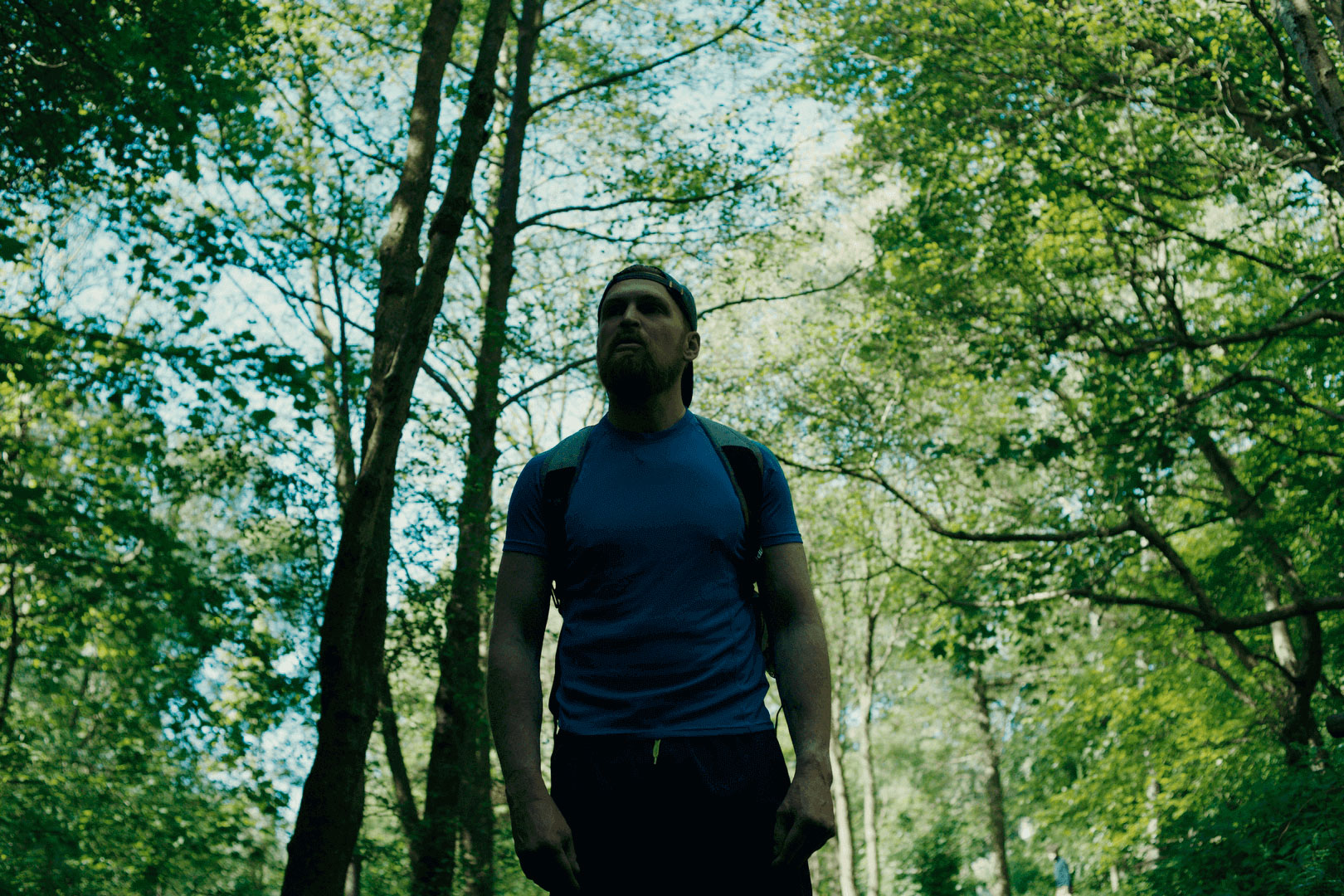 A man with a beard wearing a blue t-shirt, a backpack, and a cap turned backwards, standing in the middle of a wood.