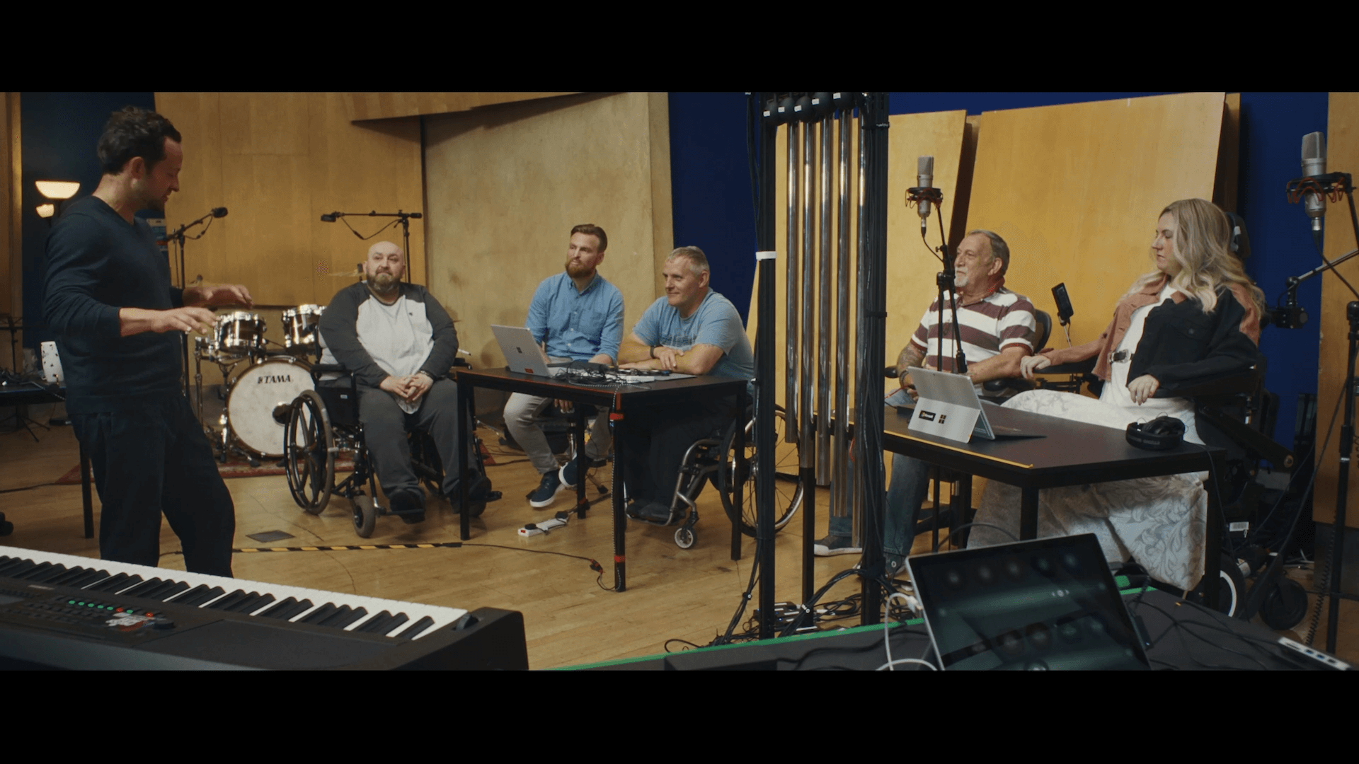 A group of people, including three in wheelchairs, are seen in a music studio looking towards a man who is standing and talking to them. The scene includes a keyboard and a set of drums in the background.