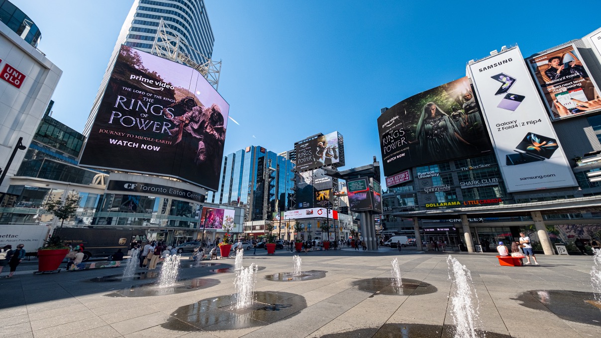 Obvious poster advertisements for Rings of Power loom above an open square with water spurting out of the floor on a sunny day with a blue sky.