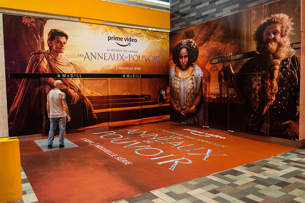 A man with grey hair stands on a large carpet poster for 'Les Anneaux De Pouvoir' or 'Rings of Power', wearing jeans and a vest top. He looks up at two large posters for the series, one featuring a dwarf with a long beard. The scene creates a sense of anticipation and excitement for the show.