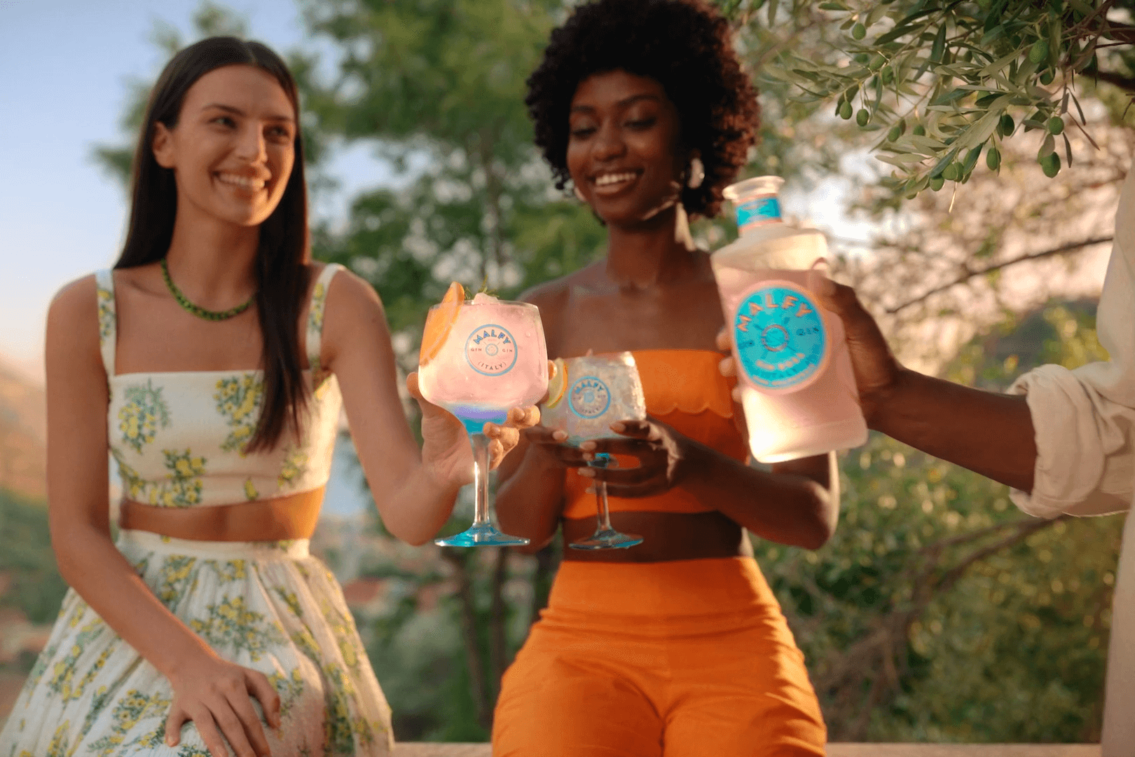 Two young women in beautiful and colorful outfits, one with an Italian flair wearing a white two-piece outfit with yellow flowers, and the other wearing an orange two-piece outfit. They stand in front of trees, holding large glasses filled with a refreshing drink with ice. A man holds a bottle of MALFY, and everyone is happy and smiling.