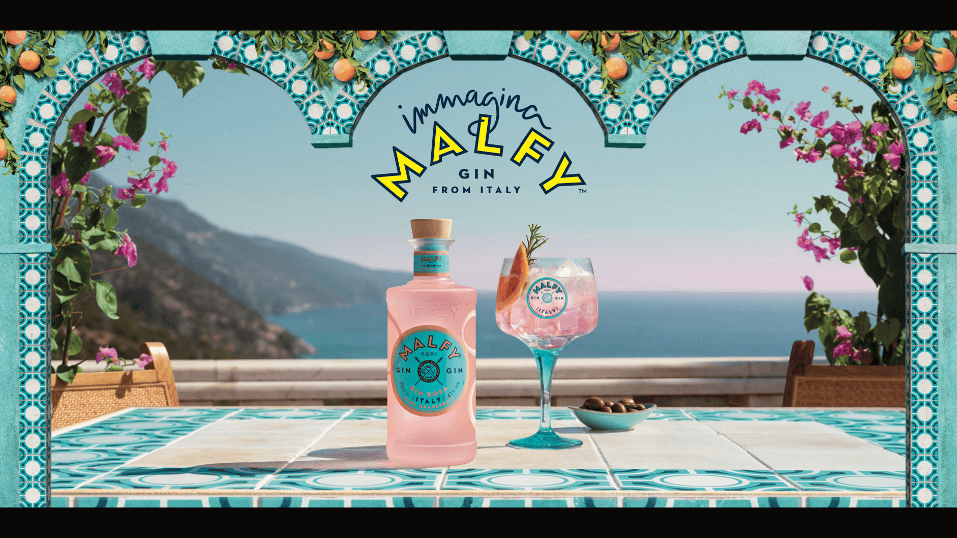 A bottle of pink Malfy Gin is prominently displayed on a table, set against a coastal backdrop. A refreshing glass, filled with ice and garnished with a sprig of rosemary, sits invitingly next to the bottle. The scene is beautifully framed by Mediterranean blue and white tiles, adding to the inviting and refreshing vibe of the image.