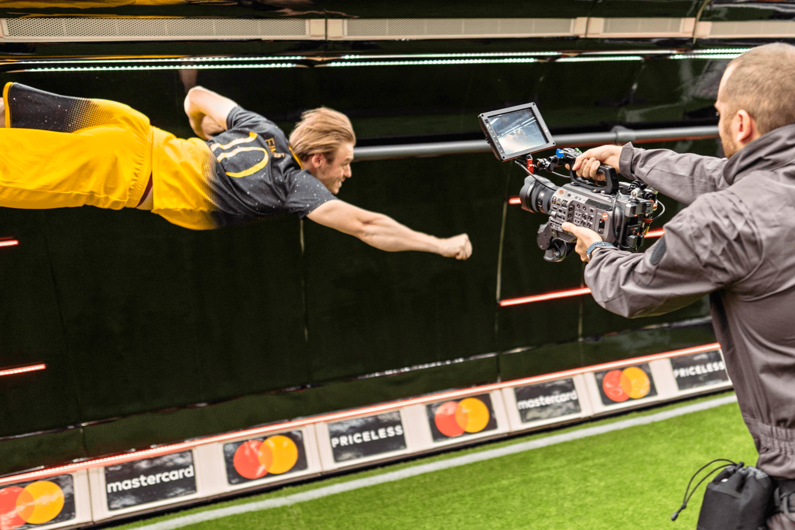 A man wearing a yellow and black football kit appears to be soaring through the air like Superman in front of a cameraman, with a Mastercard banner ad visible in the background.