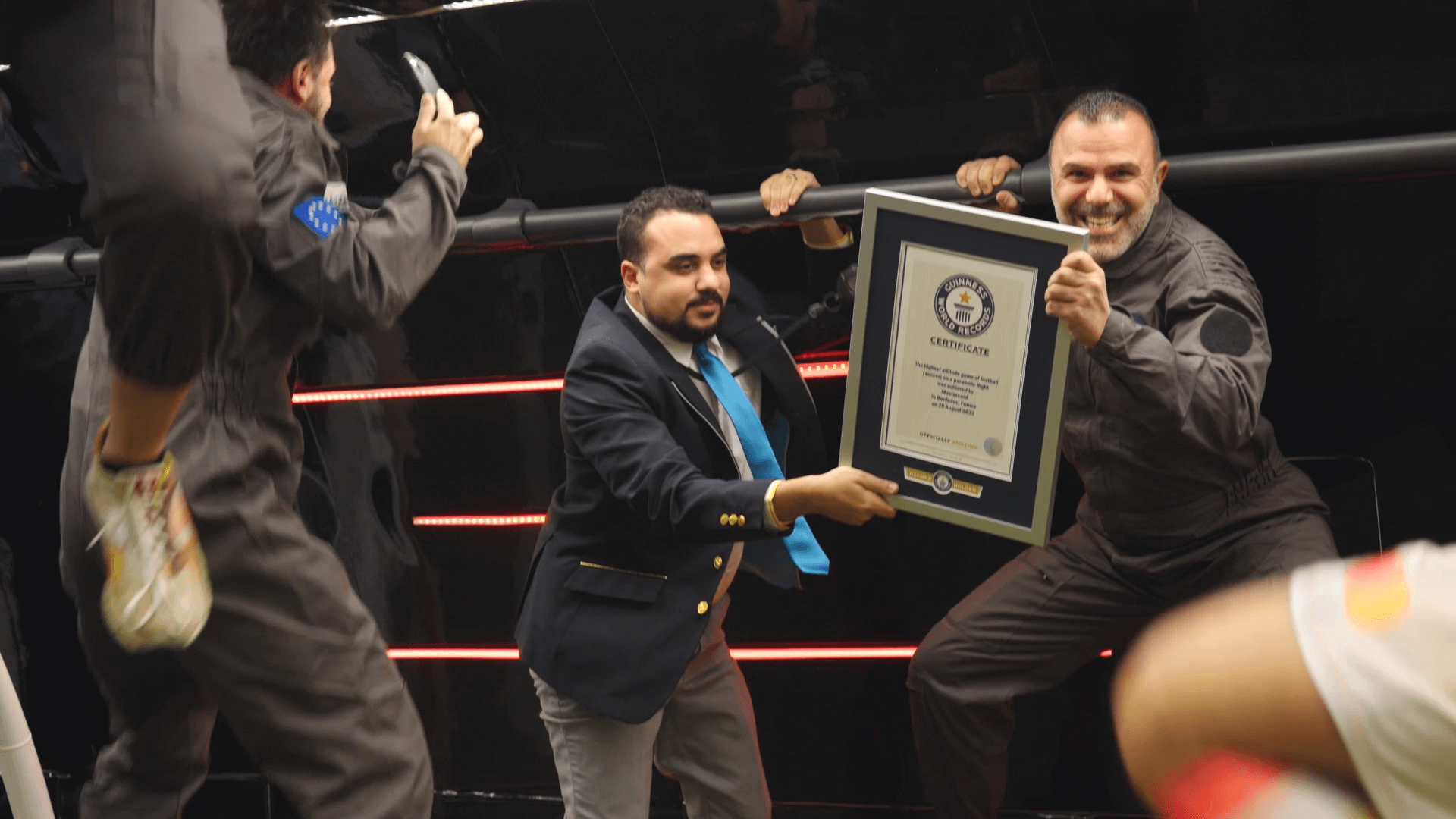 A man with a tan complexion and a mustache wearing a jumpsuit grins widely while an official hands him a framed certificate from the Guinness World Records. There are a couple of other people in jumpsuits in the foreground.