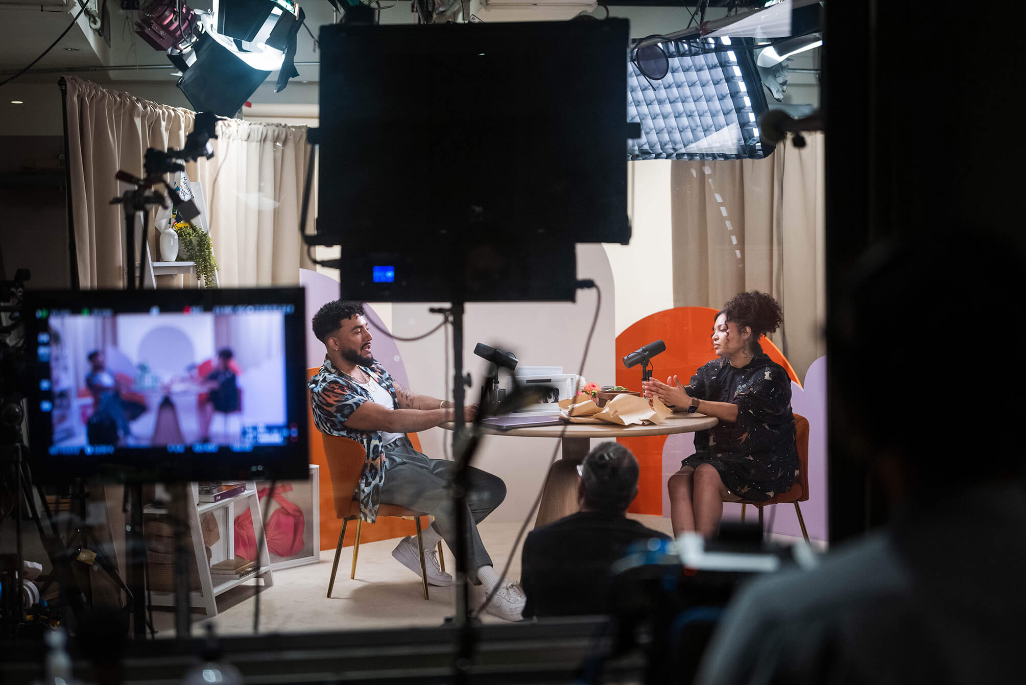 Behind the scenes, the image captures beauty expert David Lopez chatting with a stylishly dressed black lady on set. Her hair is up and she wears a black dress. Dylan wears an open graphic shirt, grey chinos, and white trainers. 