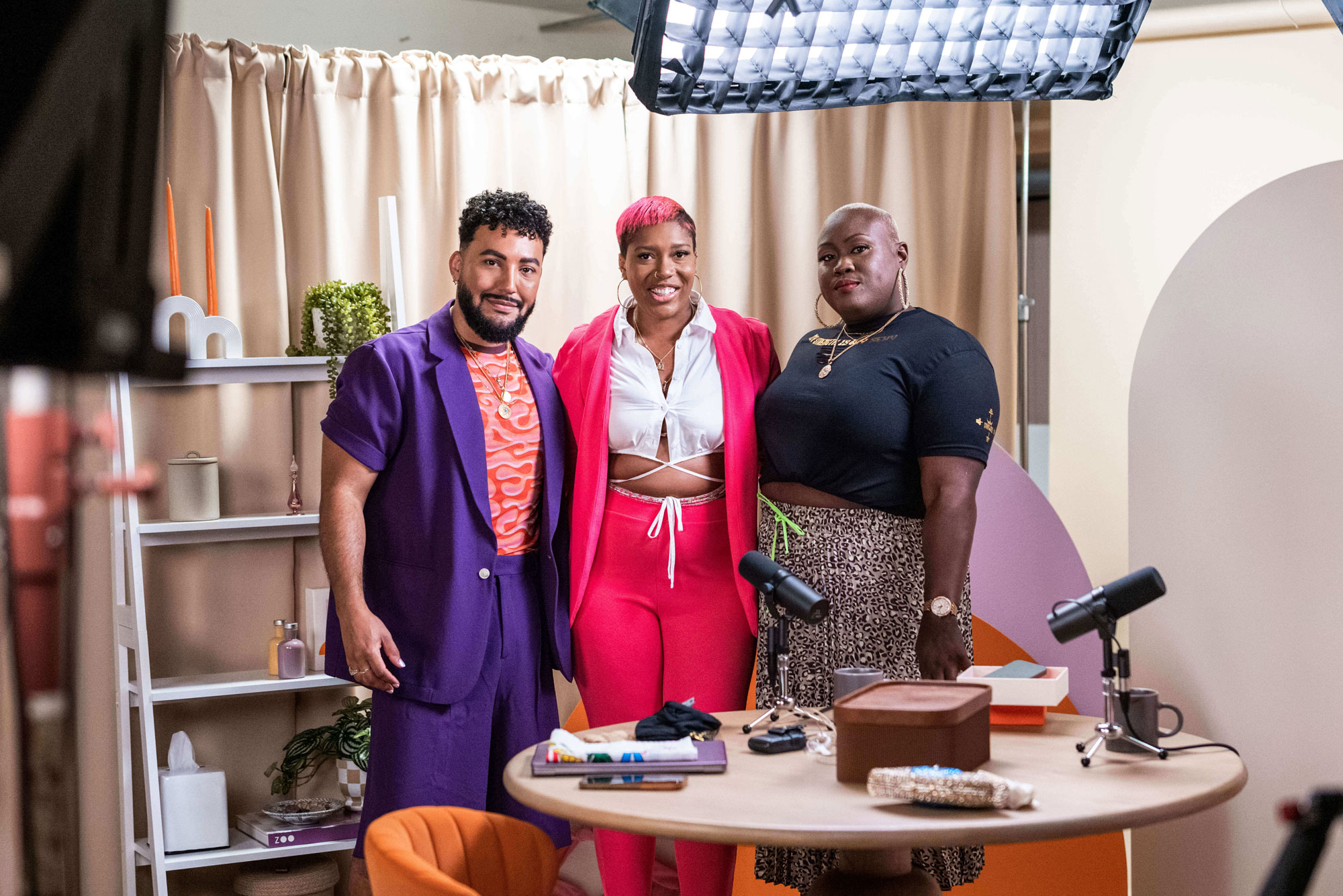 Beauty expert David Lopez poses for a picture with two striking black women. One woman is wearing a pink suit and has short pink hair, while the other has a bald head and large hoop earrings, dressed in a black outfit. David is seen in a purple suit, looking stylish and confident.