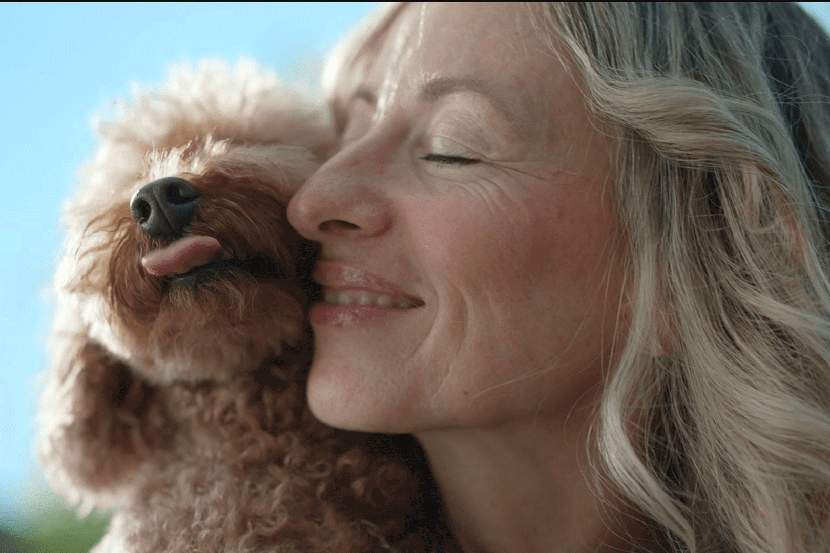 A woman with blonde hair and her eyes closed snuggles a small brown dog.