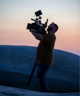 A man is holding a Steadicam camera in an open area during sunset.