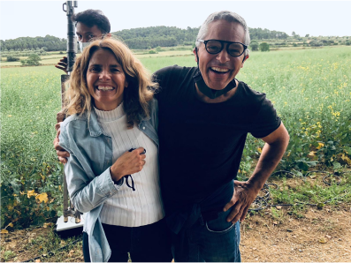 A middle aged man and woman laughing and smiling with their arms around each other in front of an overgrown field. They evoke a joyful feeling. 