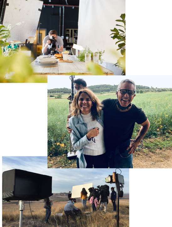 A cameraman is shooting the top of a wooden table. It has a pile of plates and a wooden board with a burger and chips. The setting is bright, but the cameraman is slightly out of focus. A middle aged man and woman laughing and smiling with their arms around each other in front of an overgrown field. They evoke a joyful feeling. A production team is in a field with light but cloudy skies overhead.