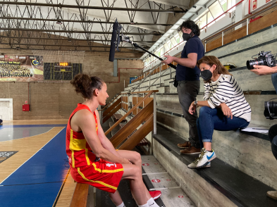 A female basketball player wearing a red and yellow uniform is being interviewed at the sideline of a court.