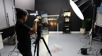 A cameraman is in a studio, filming a rack of brown bras. A large spotlight is positioned to the right.