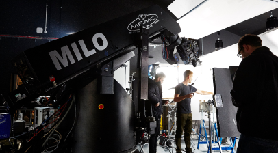 A set is being filmed behind the scenes, with a large camera crane looming over it and the team working.